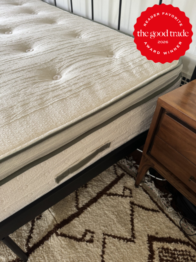 A mattress with a textured surface on a black bed frame is beside a wooden nightstand on a patterned rug. A red "The Good Trade Award Winner 2026" badge is in the top right corner.