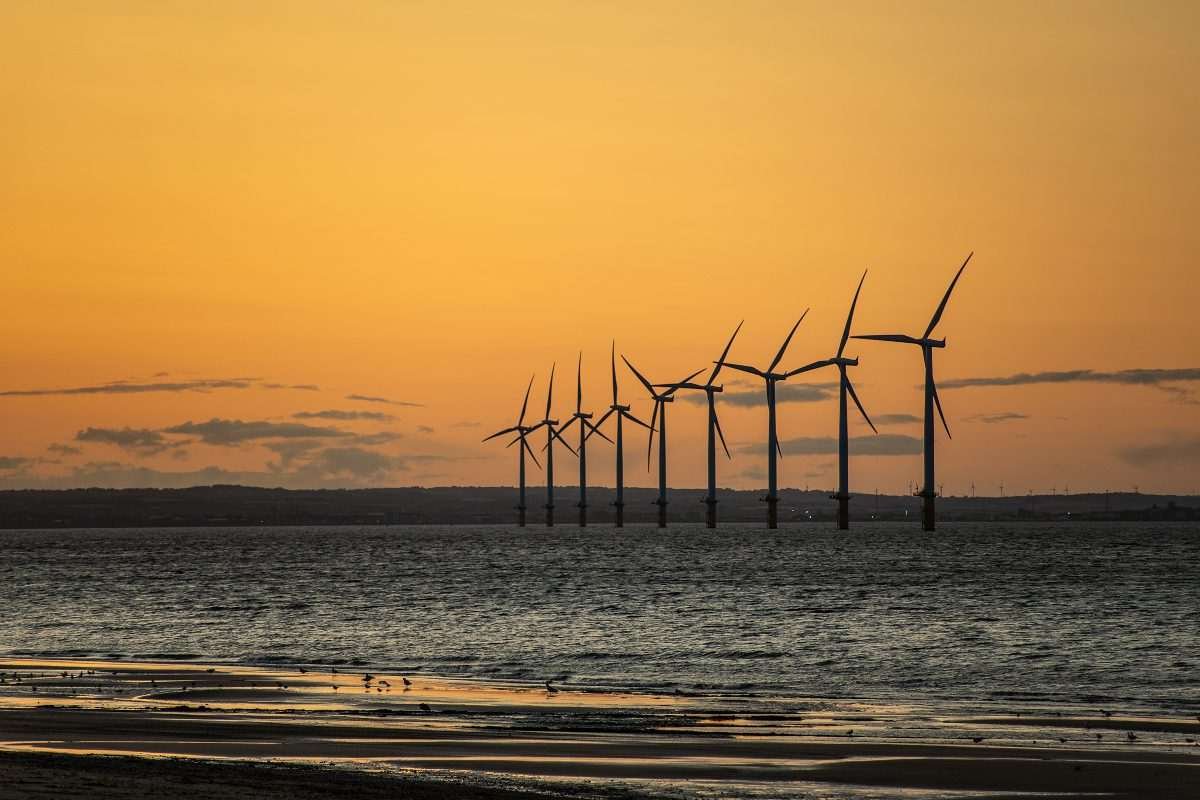 A row of offshore wind turbines in the distance, seen from the shoreline, visible as silhouettes against sunset
