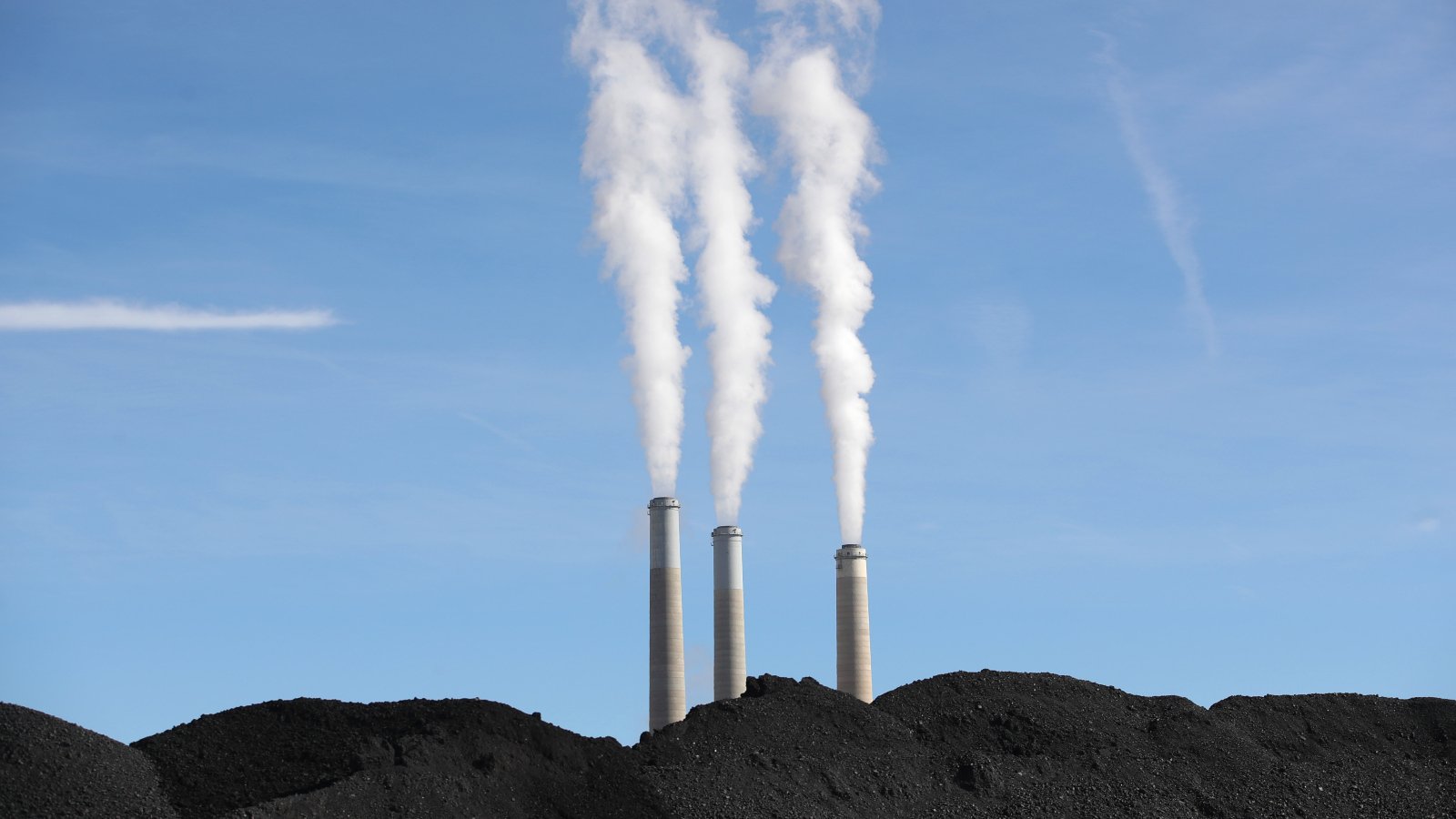 Photo of smokestacks spewing smoke into a blue sky with large mounds of coal in front