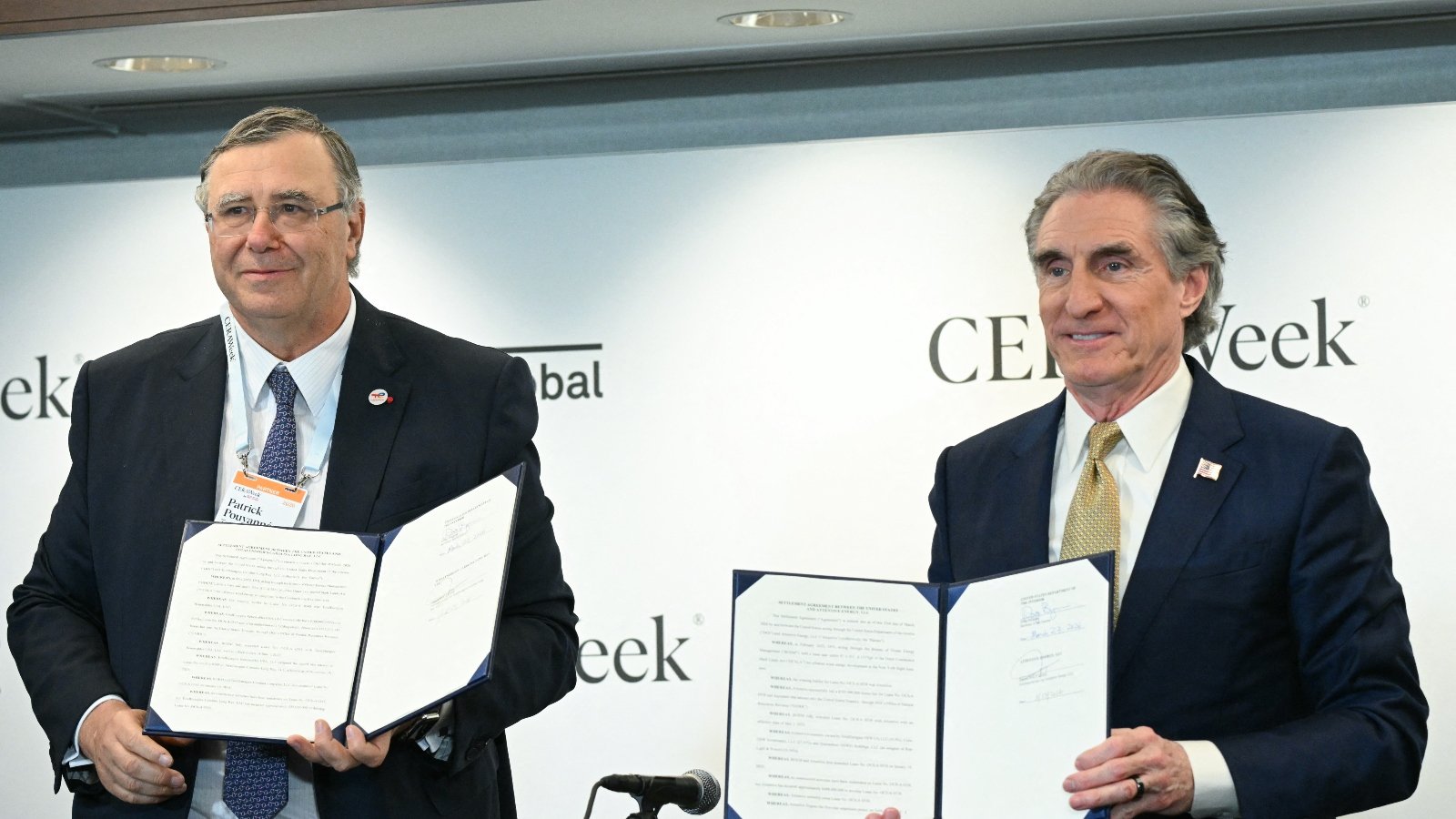Two men in business suits stand in front of a white background. They are holding papers.