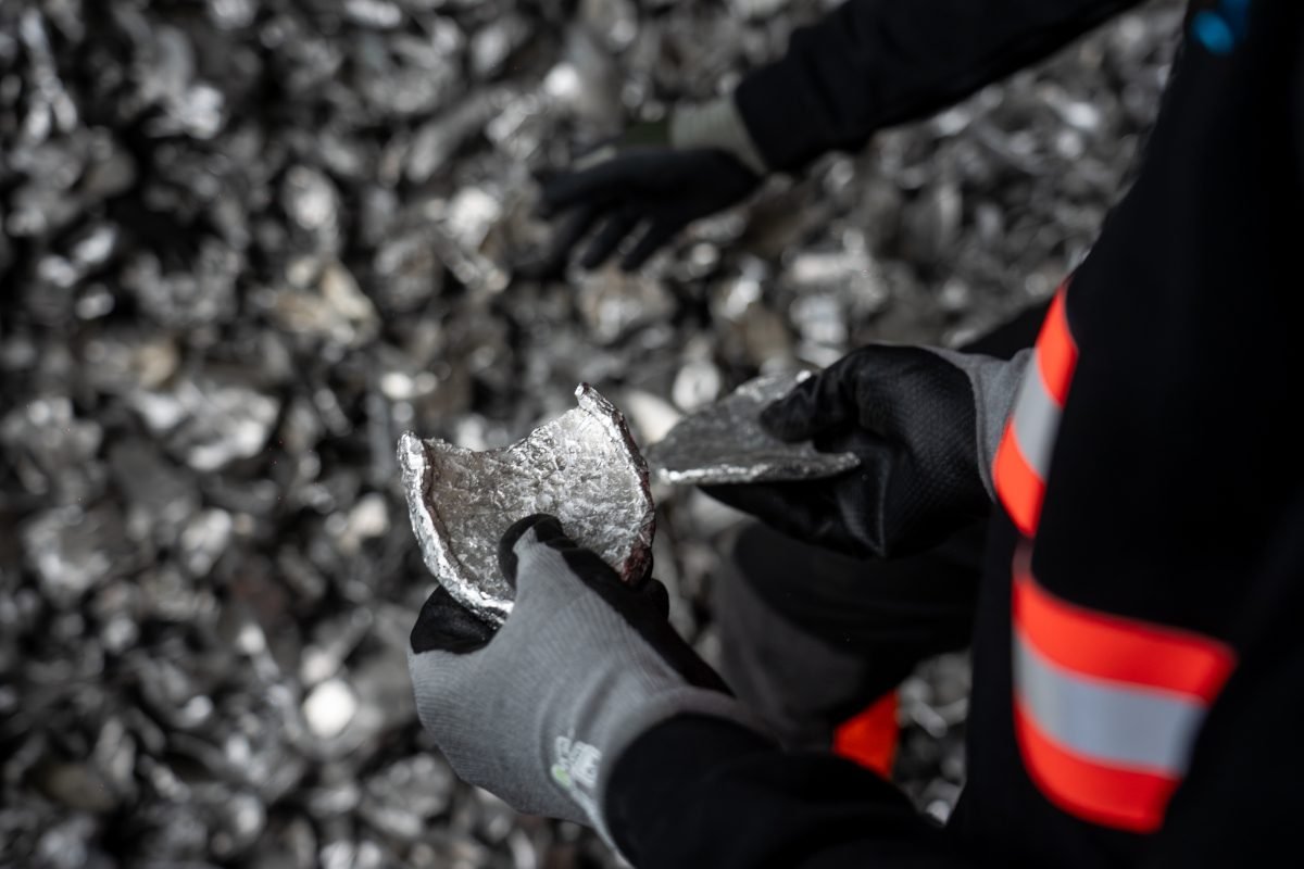 Uniformed worker's gloved hands are visible, holding a semi-circular or ovoid fragment of metal, against background of a floor entirely covered with metal fragments