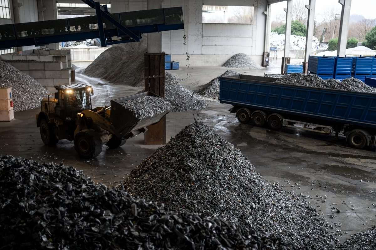 Interior industrial space showing cone-like mounds of dark or slag-like material, loader vehicles and storage vessels