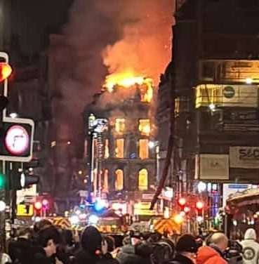 Nighttime view of a burning four-storey building with domed roof engulfed in flames, within an urban scene that includes other buildings and traffic lights nearer the viewpoint of the camera