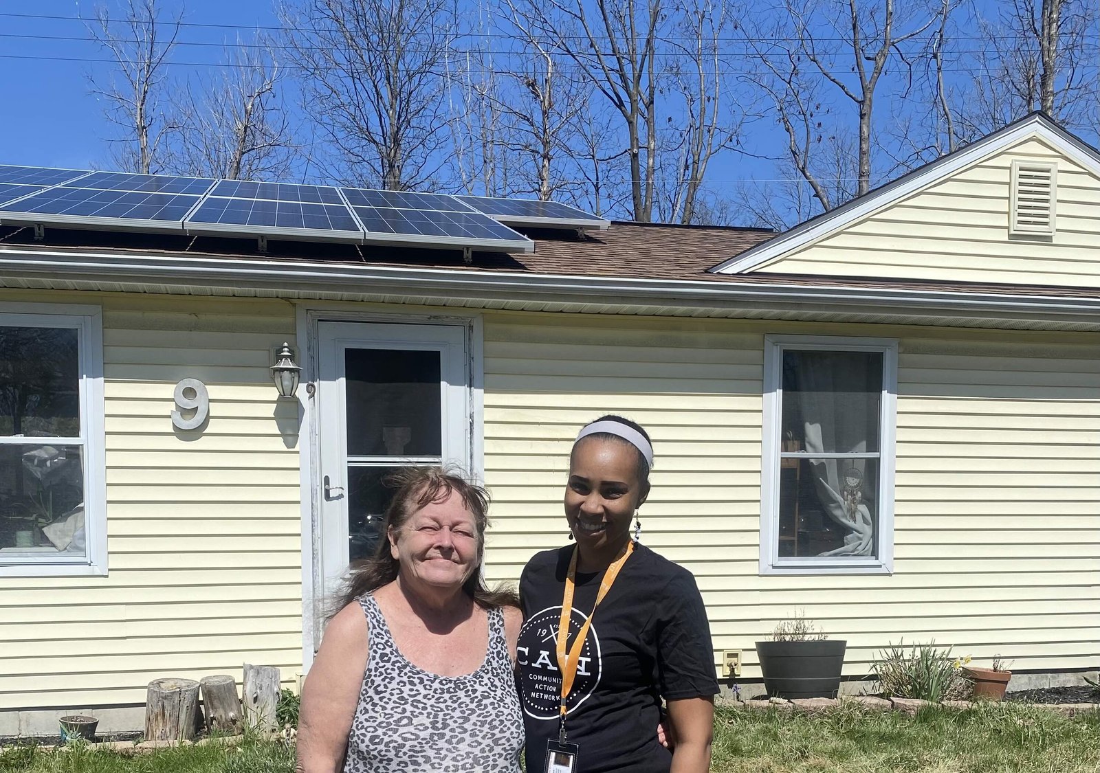 Two women stand smiling in front of a house with solar panels on its roof