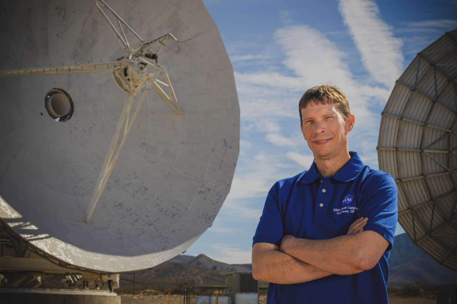 A man in a blue NASA polo shirt stands with arms crossed in front of two large satellite dish antennas at a desert ground station under a partly cloudy sky.