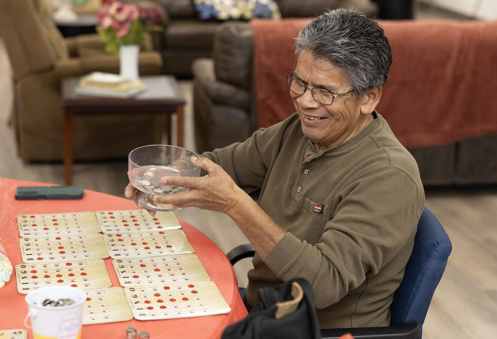 A smiling older man sits at a table in front of eight bingo cards, holding a bowl of nickels