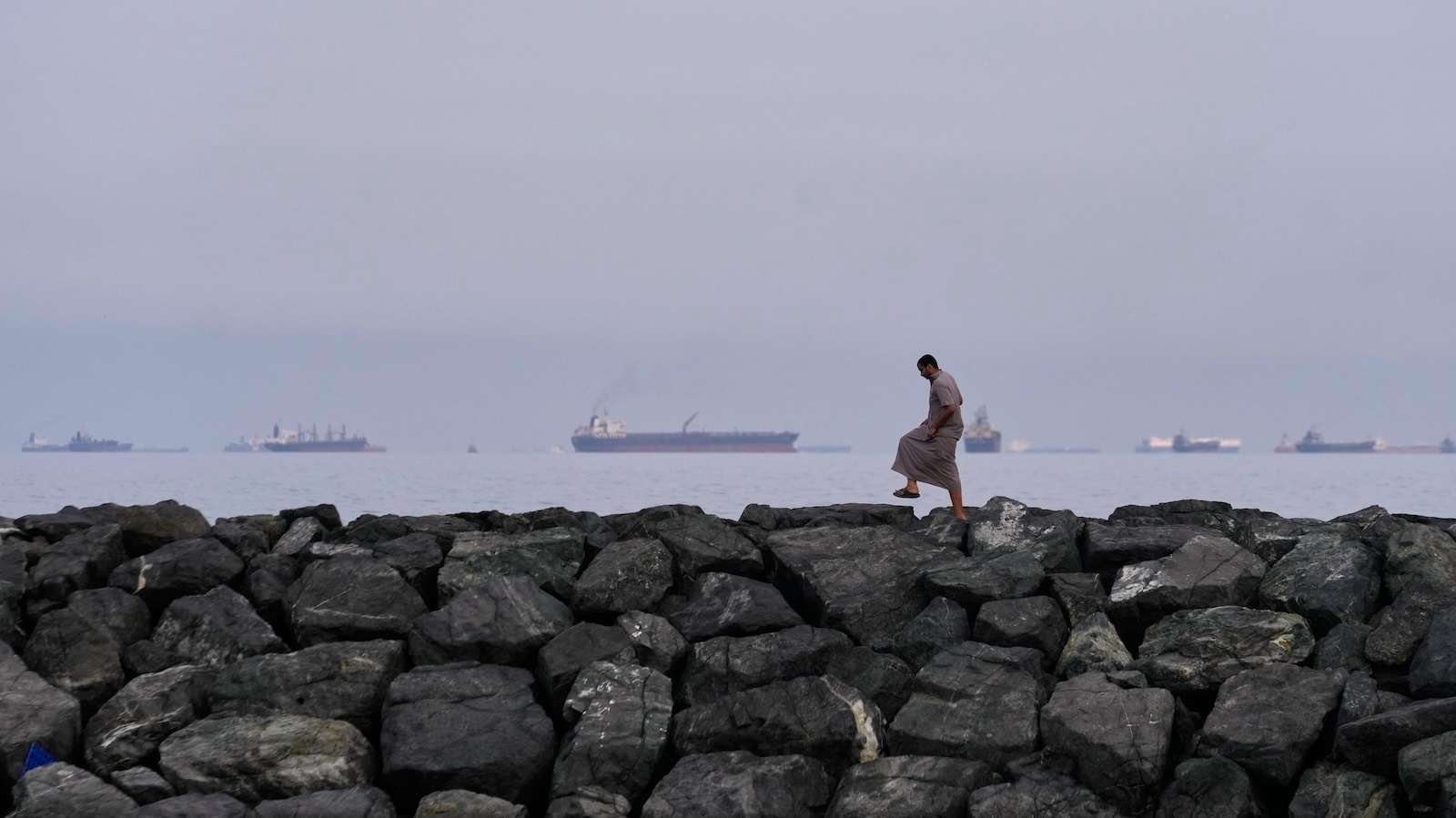 A man walks on rocks along the shore as oil tankers and cargo ships line up in the Strait of Hormuz, as seen from Khor Fakkan, United Arab Emirates, Wednesday, March 11, 2026. (AP Photo/Altaf Qadri)