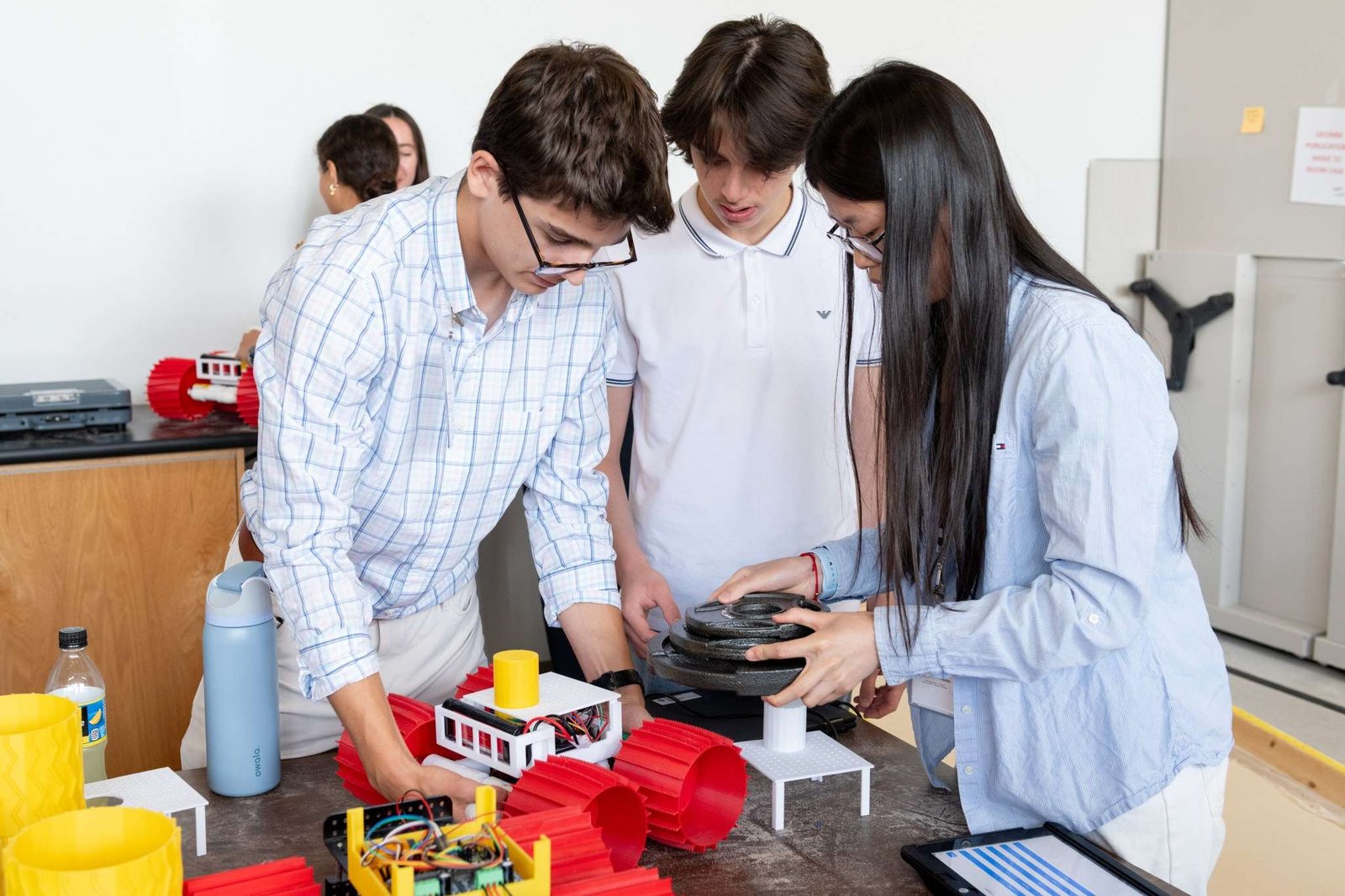Three High School Engineering Institute students are centered in the foreground of the image working on a small vehicle with large red plastic wheels. There are two more students in the background working on their own identical vehicle.