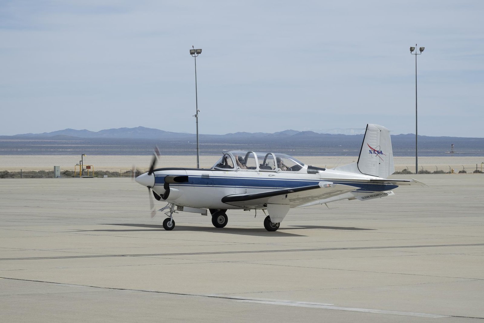 A small white aircraft with a blue stripe, and a black front propellor, drives along a concrete ramp with the desert and mountains behind it. There are two people inside, and only the tops of the helmets of both people can be seen under the clear canopy of the aircraft.