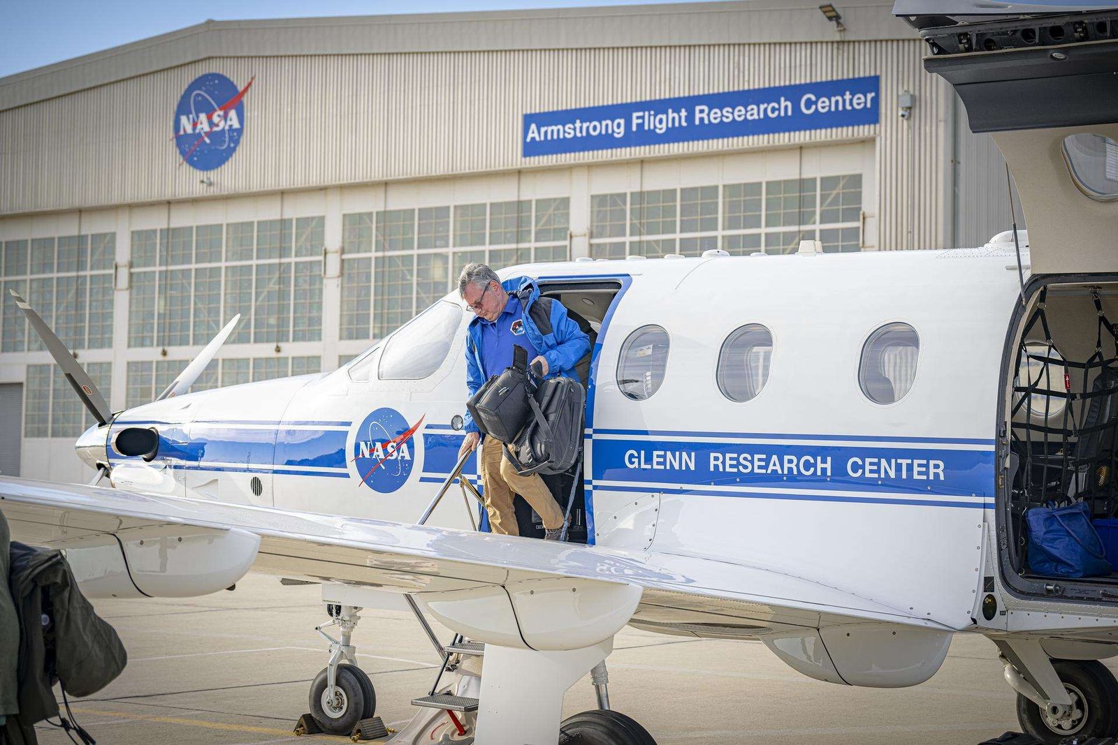 The left side of a white aircraft with one door and three windows sits on a concrete ramp. The aircraft is painted with a blue stripe and a round NASA logo on the side. A man in a blue jacket and tan pants climbs out of the aircraft carrying two black bags. A silver hangar sits in the background with large white doors. A round NASA logo is located on the hangar as well as a blue sign with white letters to the right side.
