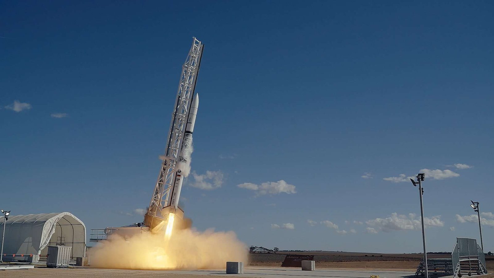 Long thin space rocket during launch, photographed midway up the launch tower and with a fiery jet and large exhaust plume also visible, all situated within a flat, remote-seeming landscape with mostly clear blue sky overhead