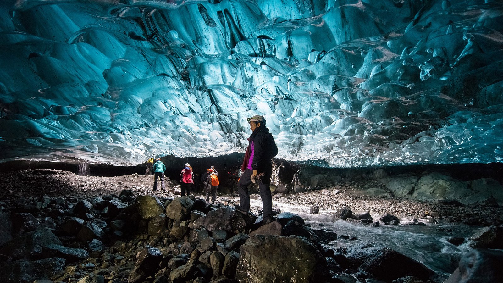 a group of tourists in coats and helmets stand inside a cave, beneath an impressive shiny blue and silver wall of ice