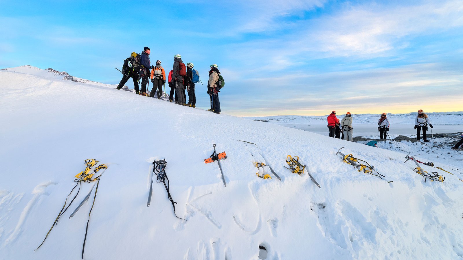 A group of tourists stands on top of a snowy slope, with their crampons lined up in the snow in the foreground