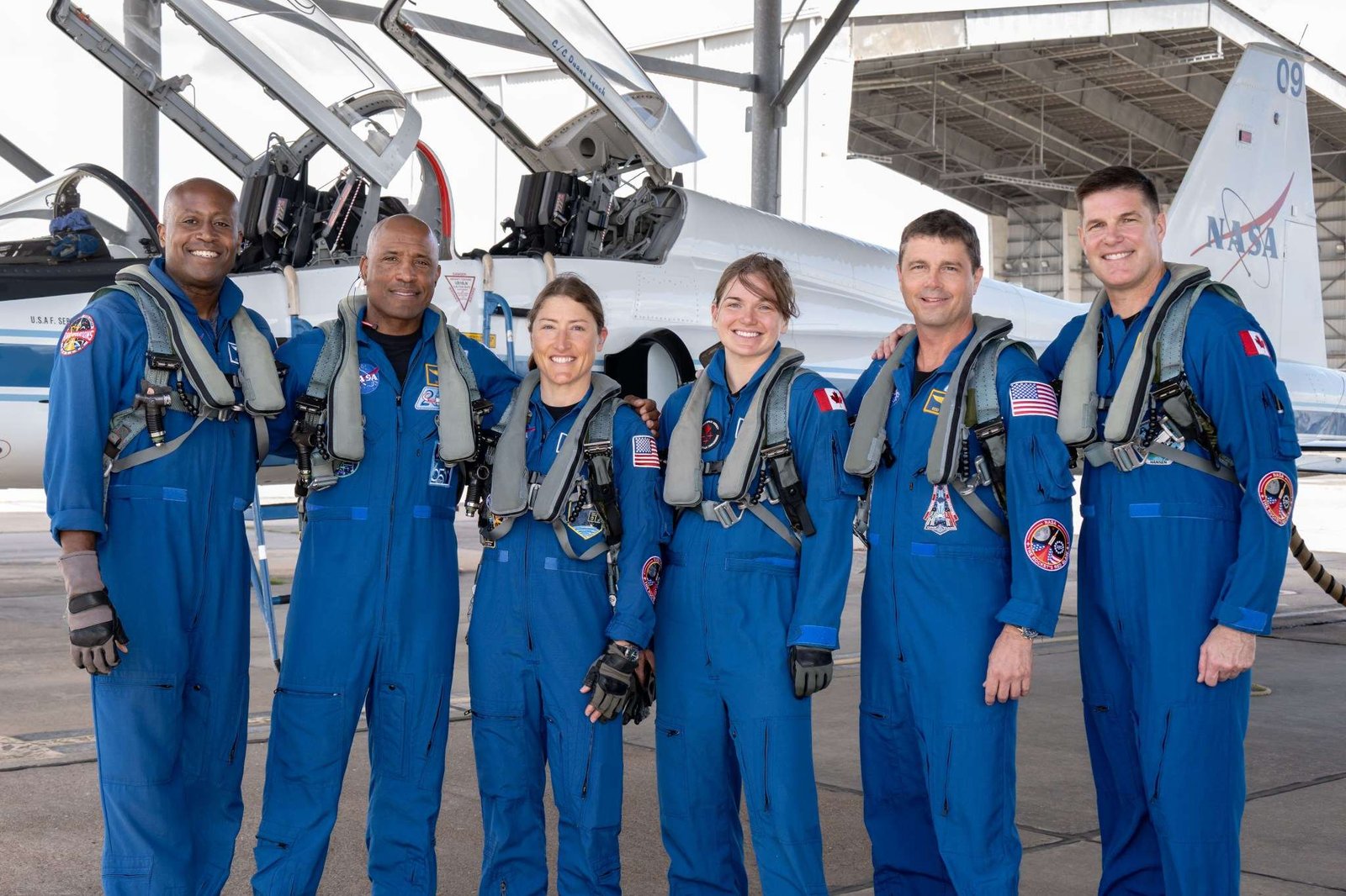 From left to right, NASA astronauts Andre Douglas, Victor Glover, and Christina Koch, CSA (Canadian Space Agency) astronauts Jenni Gibbons, NASA astronaut Reid Wiseman, and CSA astronaut Jeremy Hansen pose in front of an airplane. They are all wearing blue jumpsuits with patches and gray harnesses.