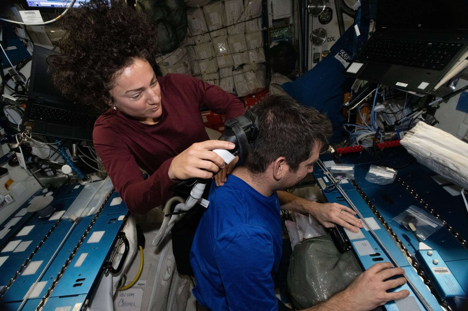 Jessica Meir holds an electric razor with a vacuum to Jack Hathaway's hair. She looks down at what she's doing, while Hathaway faces to the right of the image. Meir's brown curly hair floats up above her head.