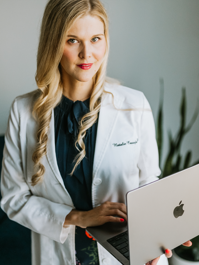 Person in a white coat holding an open laptop, standing indoors with a plant in the background.