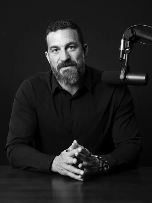 Man with a beard sits at a table in front of a microphone against a dark background.