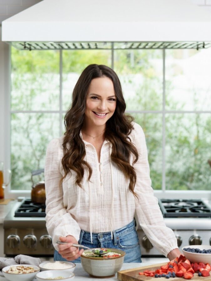 A person in a kitchen prepares food, standing near a stove with bowls of strawberries, blueberries, and nuts on the counter.