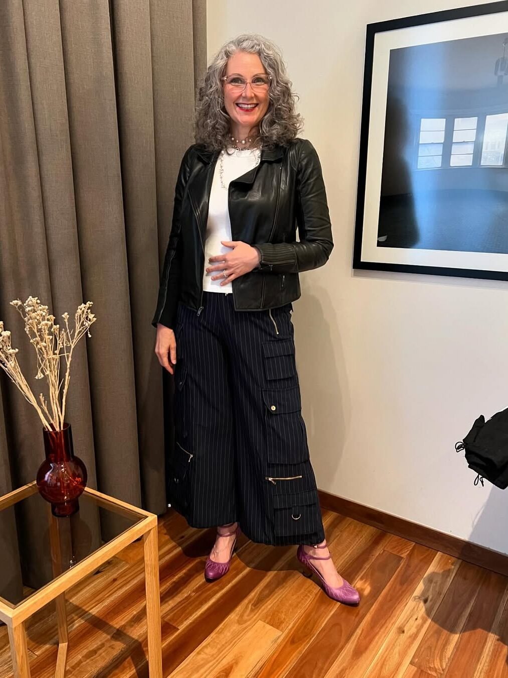 A woman with gray hair, wearing a black leather jacket, white top, and striped pants, stands in a room with wooden flooring and a framed picture on the wall.