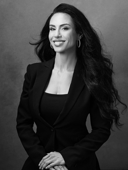 Woman with long hair and hoop earrings, wearing a suit, smiles at the camera in a black and white portrait.