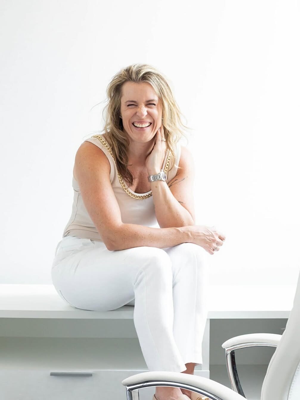 A woman with blonde hair sits on a desk, smiling, wearing a sleeveless top and white pants. A white office chair is partially visible in the foreground.