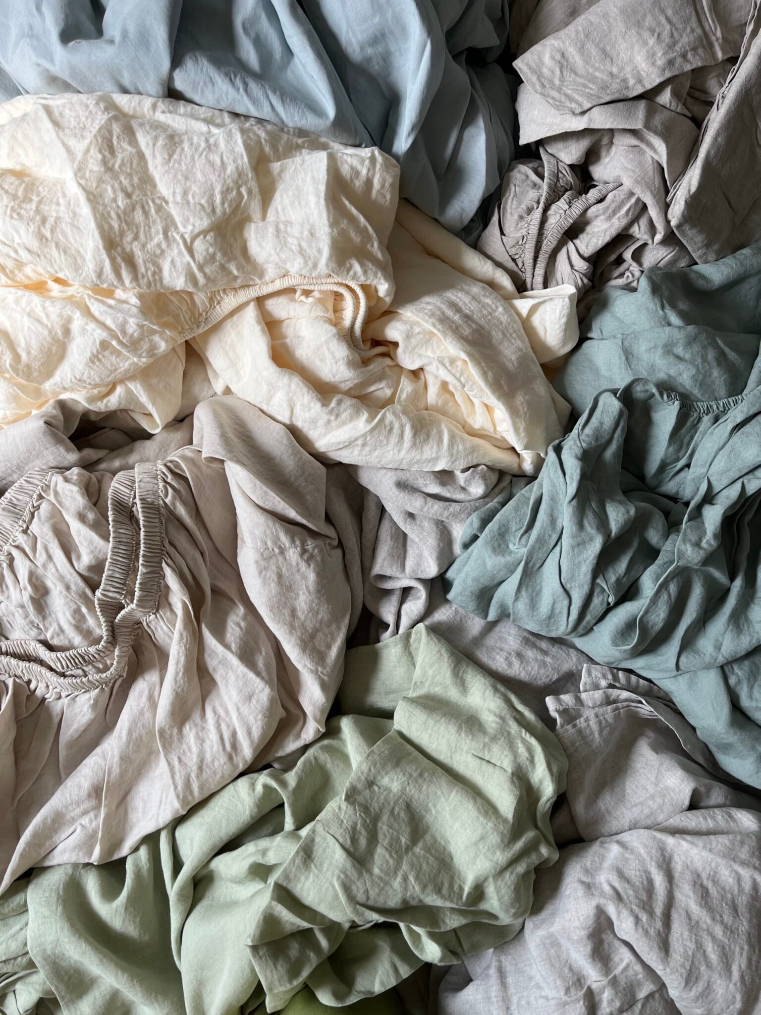 A jumble of wrinkled bedsheets and linens in various muted colors, including beige, light blue, green, and cream, spread out closely together.