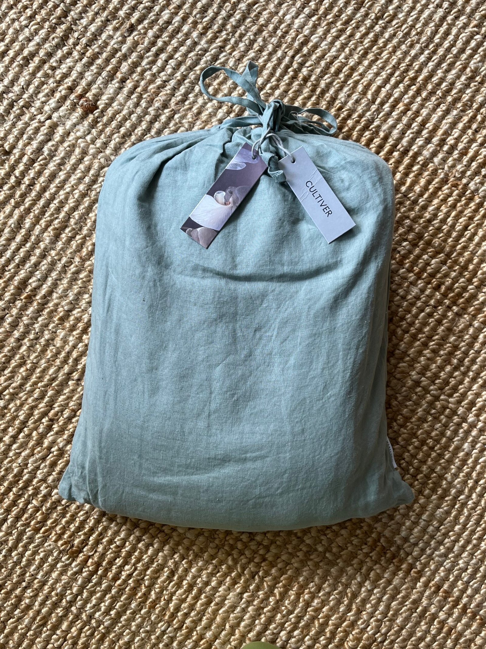 A light green fabric bag tied with a string, labeled "Cultiver," placed on a woven jute rug.