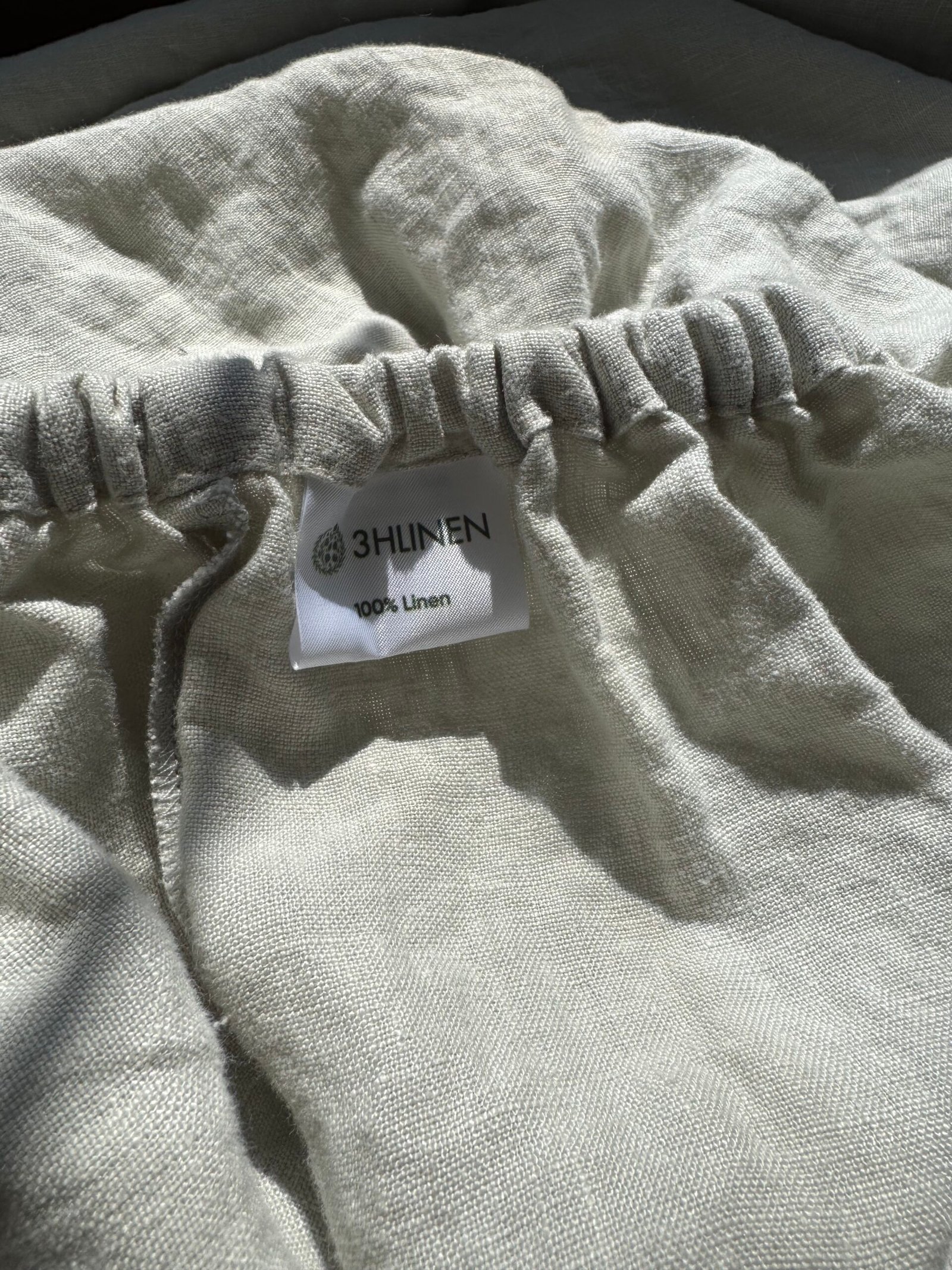 Close-up of a linen fabric with a gathered elastic band and a label that reads "3HLINEN 100% Linen." The fabric appears to be light and textured.