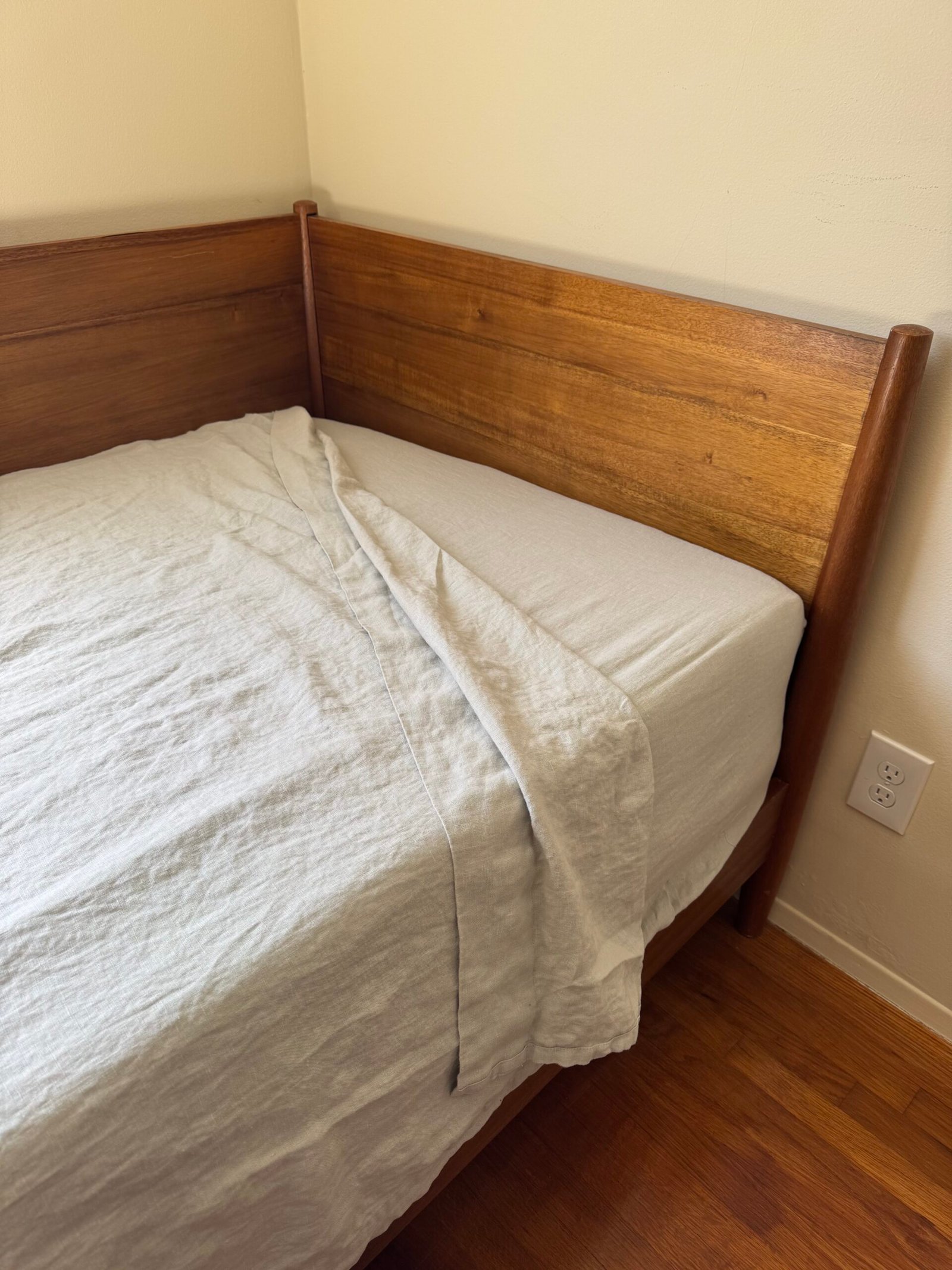 Wooden bed frame with a light gray fitted sheet and blanket partially folded back, set on a hardwood floor near a corner wall and electrical outlet.