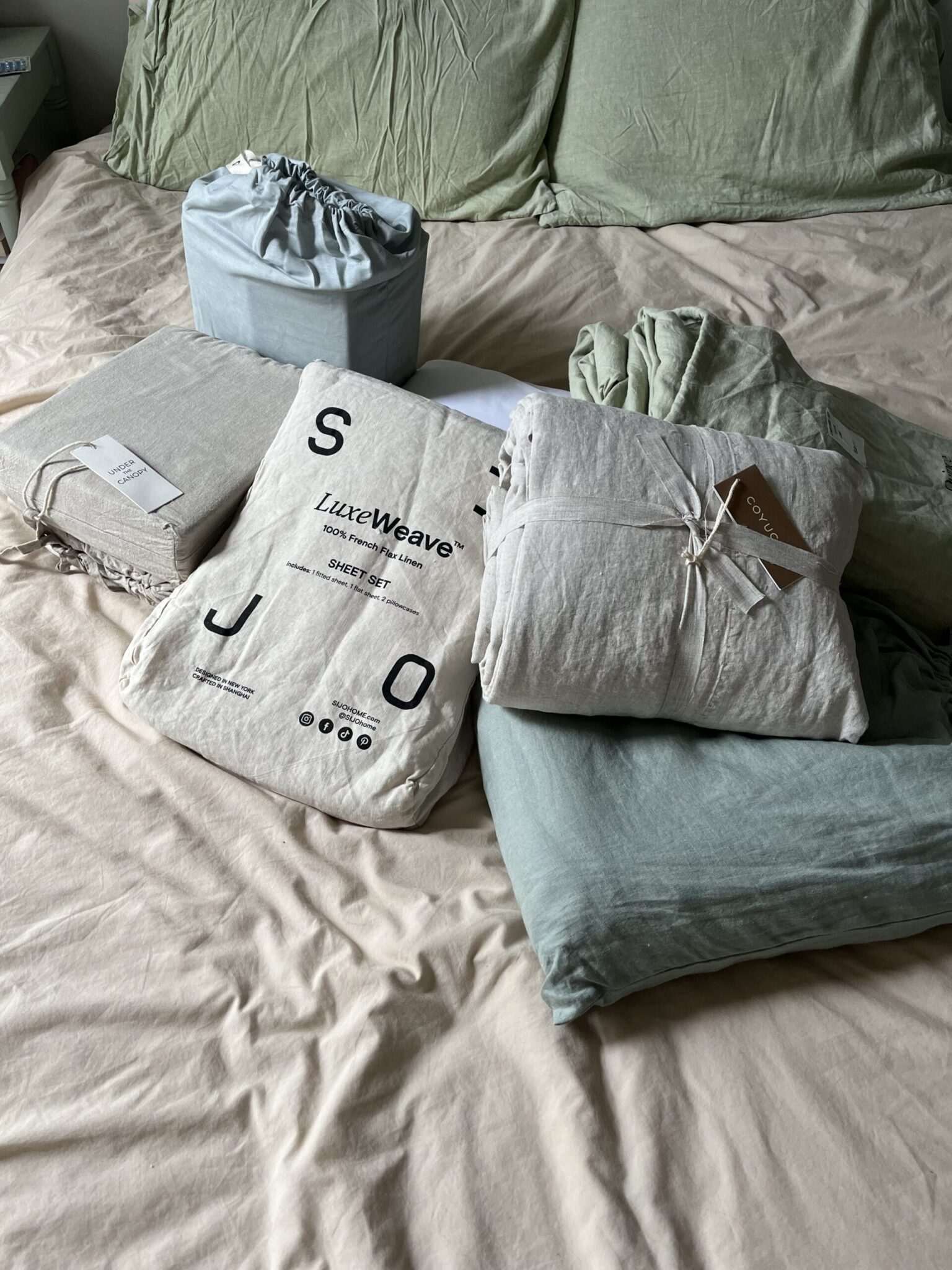 A variety of packaged bedding items, including sheet sets and pillowcases in green and beige tones, are arranged on a made bed with green pillows.