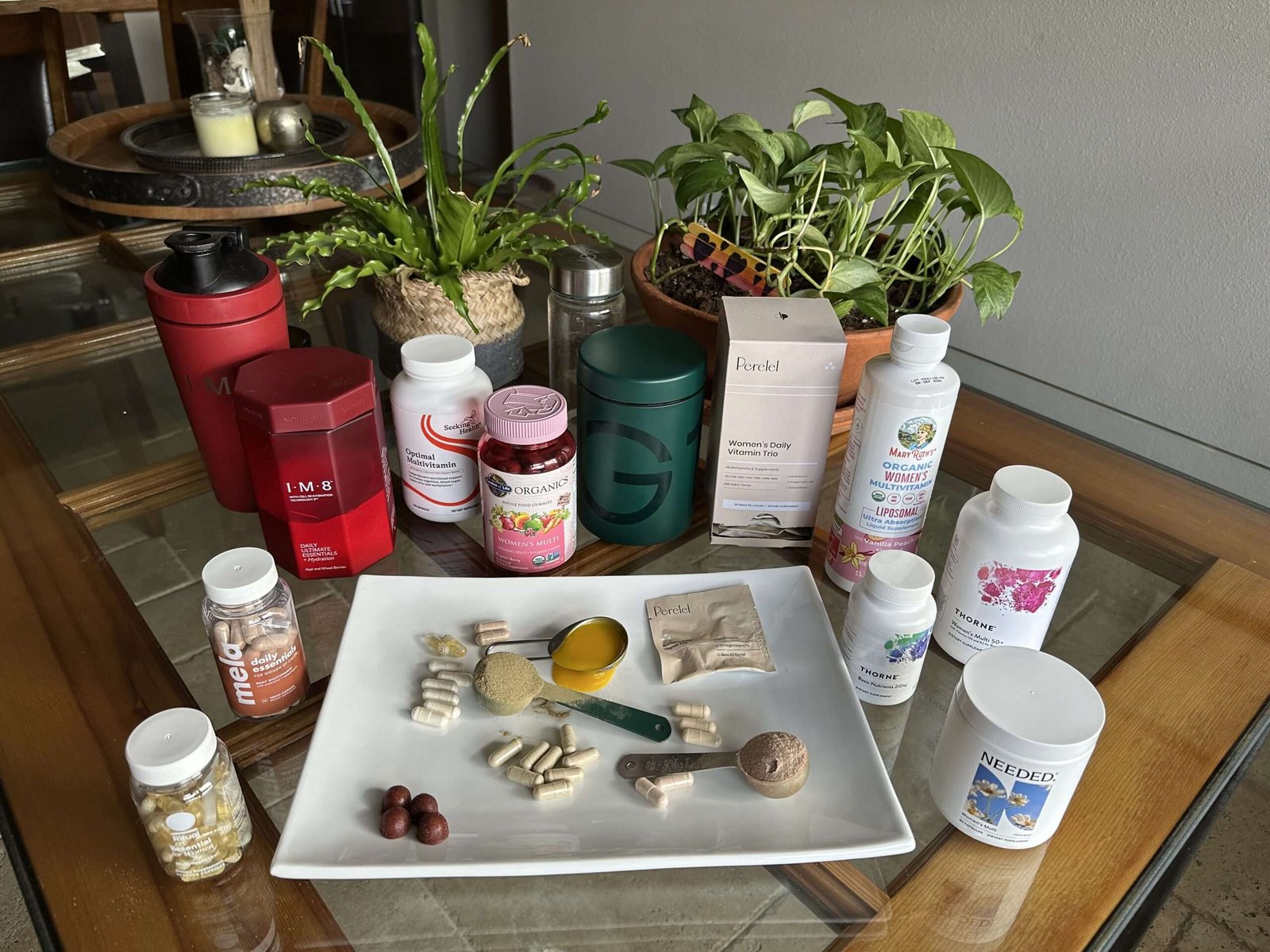 A variety of supplement bottles, capsules, powders, and drinks are arranged on a wooden table with green plants in the background.