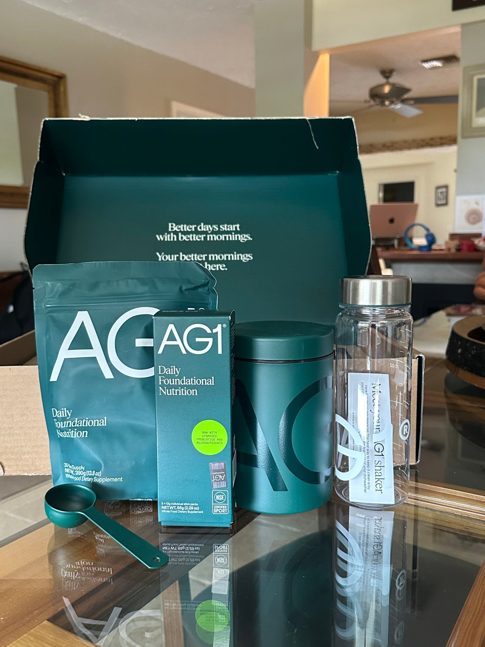 A box of AG1 nutrition products on a table, including a pouch, a bottle, a shaker, a scoop, and a canister, with the box lid displaying a motivational message.