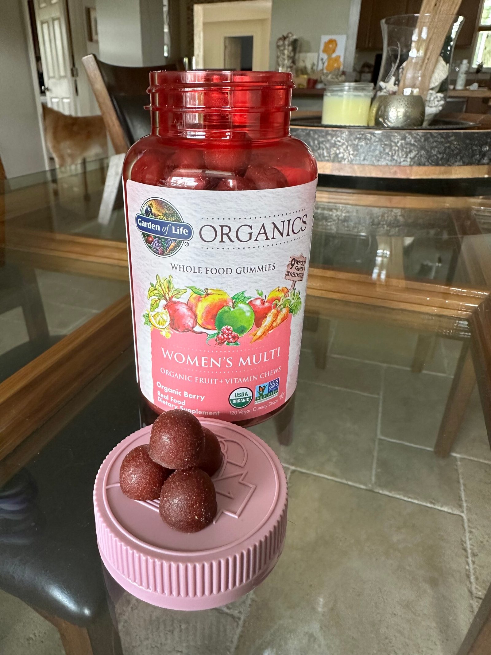 A bottle of Garden of Life Organics Women's Multi whole food gummies with several gummies placed on the lid, on a kitchen table.