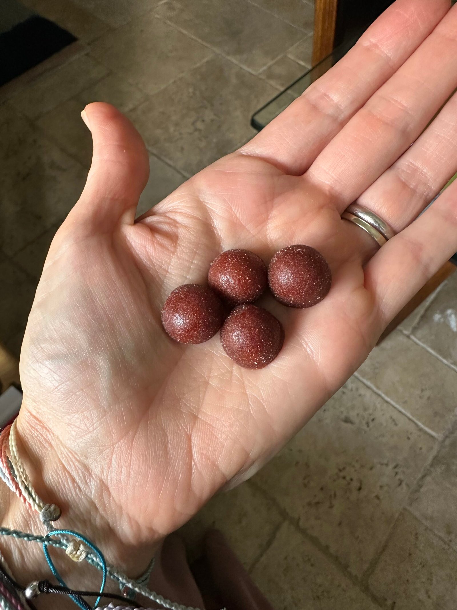 A hand holding four round, reddish-brown, sugar-coated candies or gummies over a tiled floor.