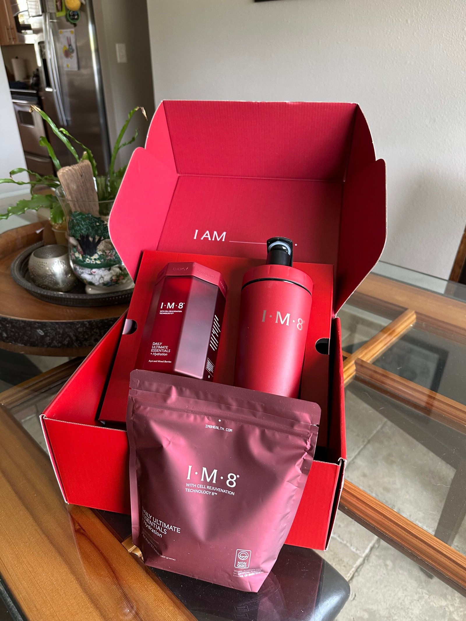 A red box labeled "I AM 8" contains a bottle, a canister, and a pouch of nutritional supplement products on a glass table next to a potted plant.
