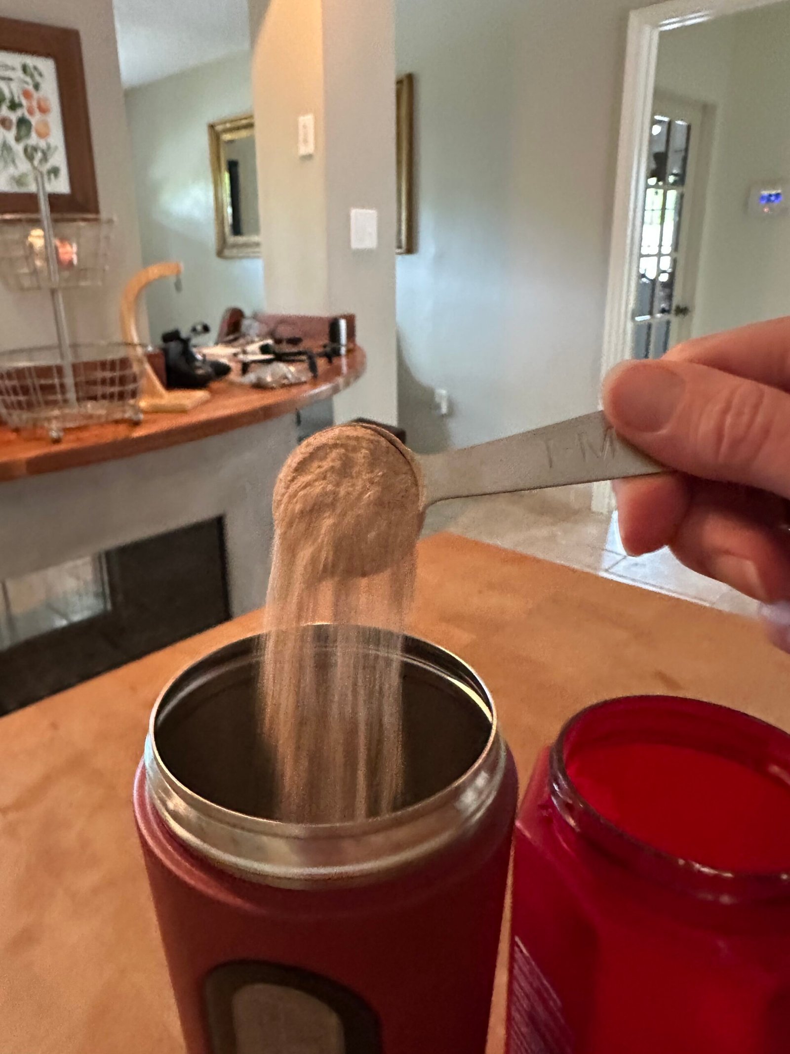 A hand holds a tablespoon, pouring powder into a red container on a kitchen counter with household items in the background.
