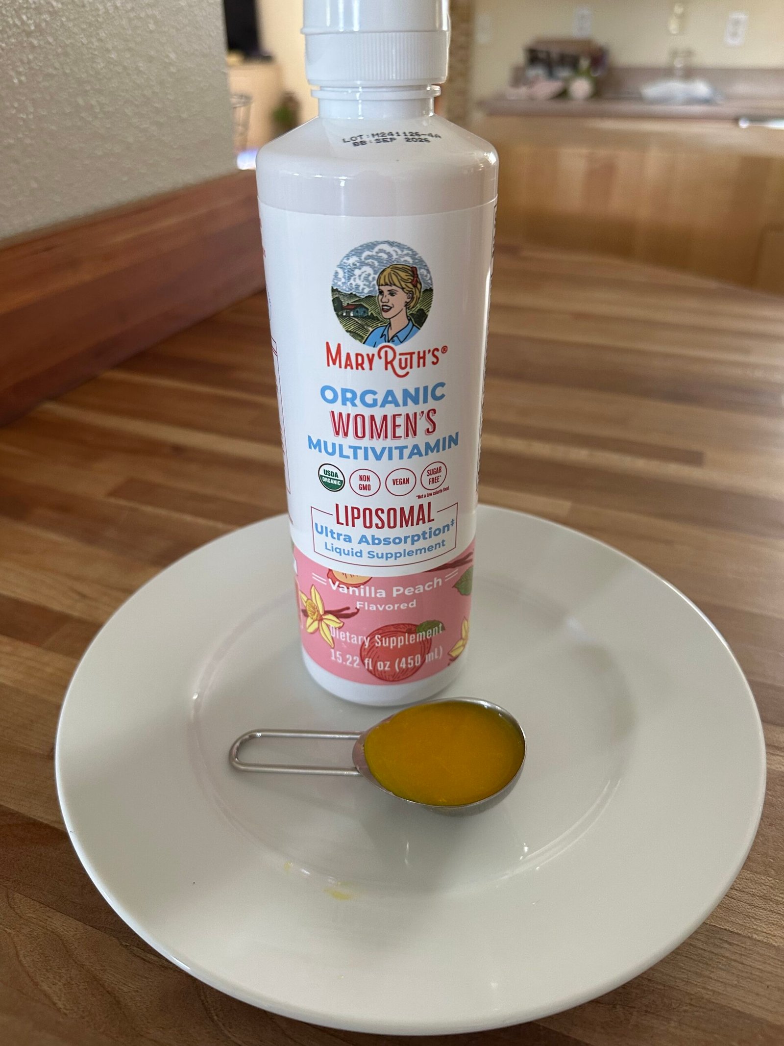 A bottle of MaryRuth's Organic Women's Multivitamin stands on a plate, with a metal tablespoon filled with the liquid vitamin in front.