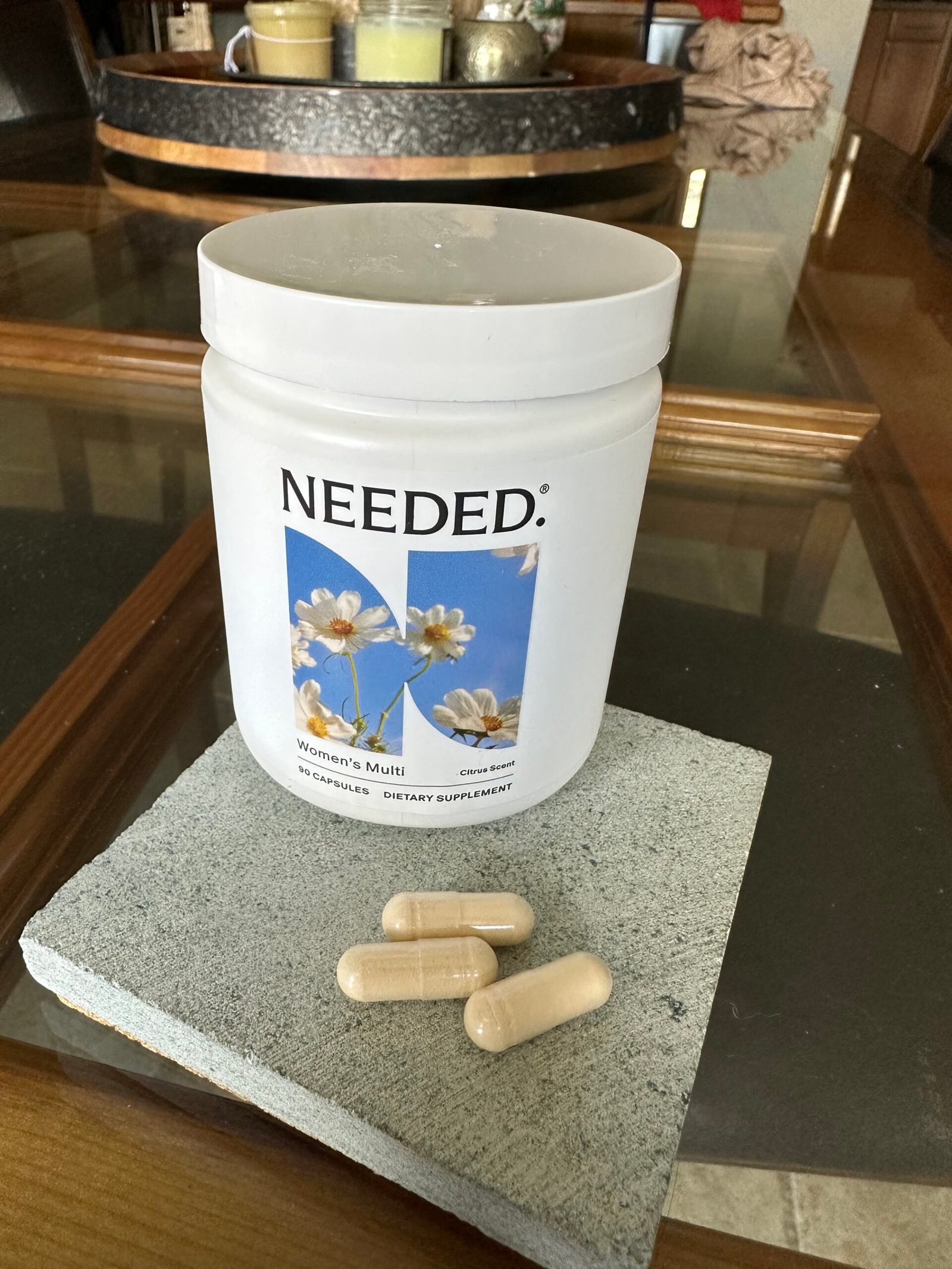 A container labeled "Needed: Women's Multi" sits on a coaster with three beige capsules next to it, on a glass table with candles and decor in the background.