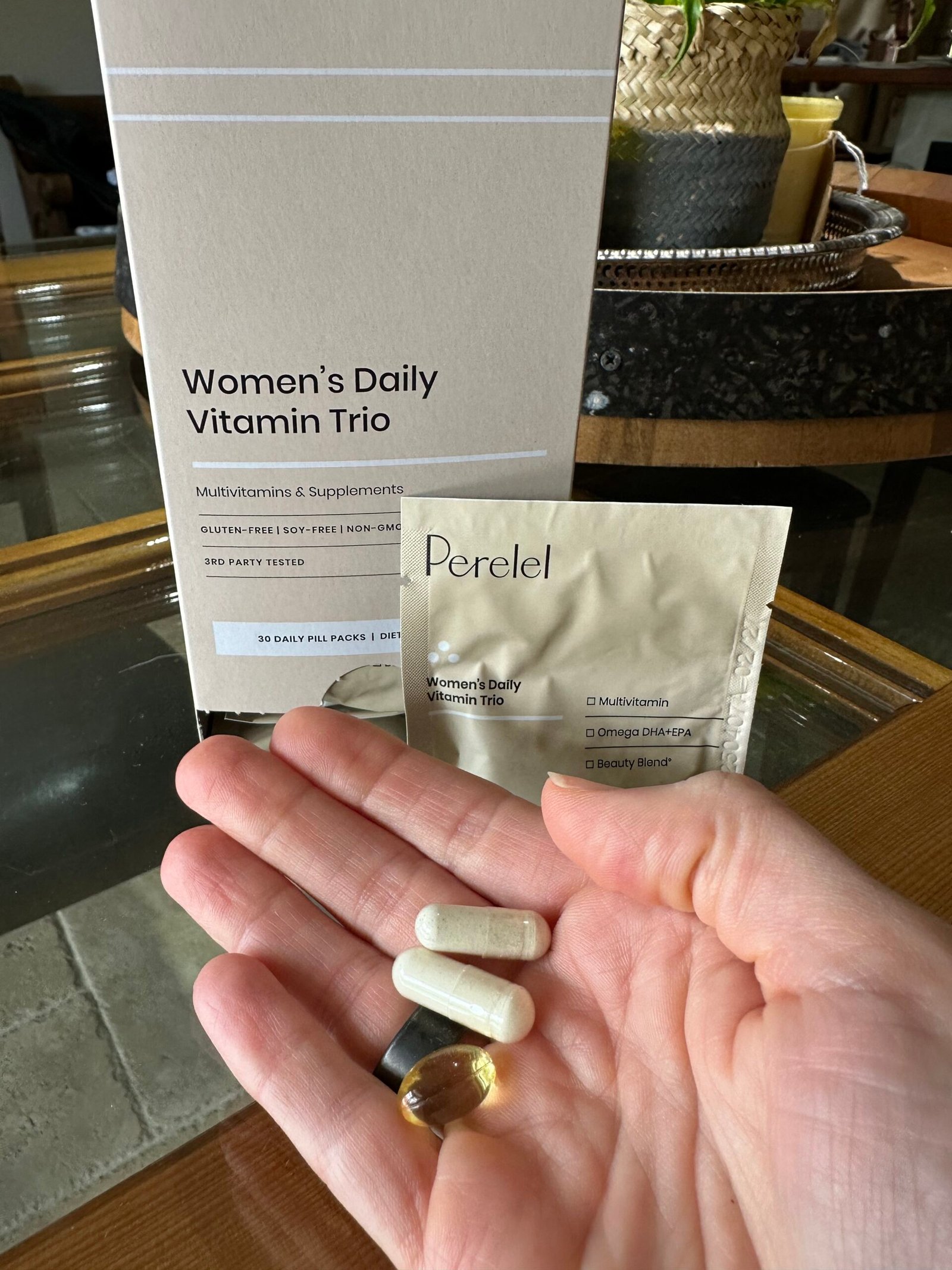 A hand holds four supplement pills in front of a Perel Women's Daily Vitamin Trio box and a packet labeled with the same product name.