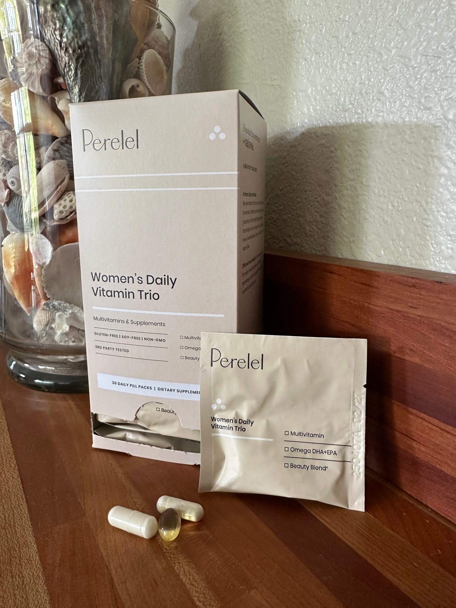 Box and packet of Perelel Women's Daily Vitamin Trio on a wooden surface, with three supplement capsules and a glass container of seashells in the background.