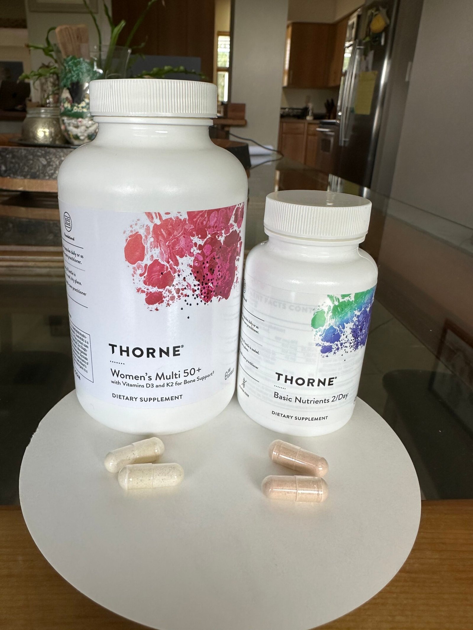 Two bottles of Thorne dietary supplements, Women's Multi 50+ and Basic Nutrients 2/Day, are placed on a table with five assorted capsules in front of them.
