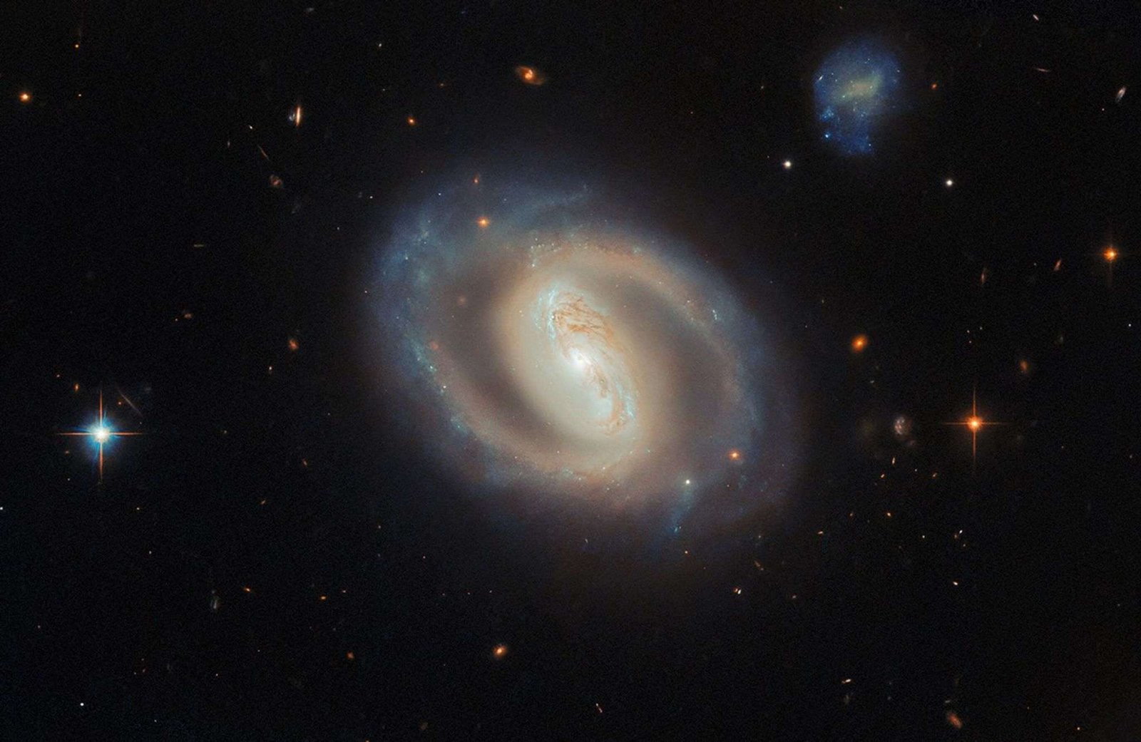 A face-on view of the barred spiral galaxy IC 486, showing a bright, elongated central bar and softly curving, ring-like spiral arms with subtle blue star-forming regions and dark dust lanes, set against a black background dotted with distant galaxies and a few foreground stars.
