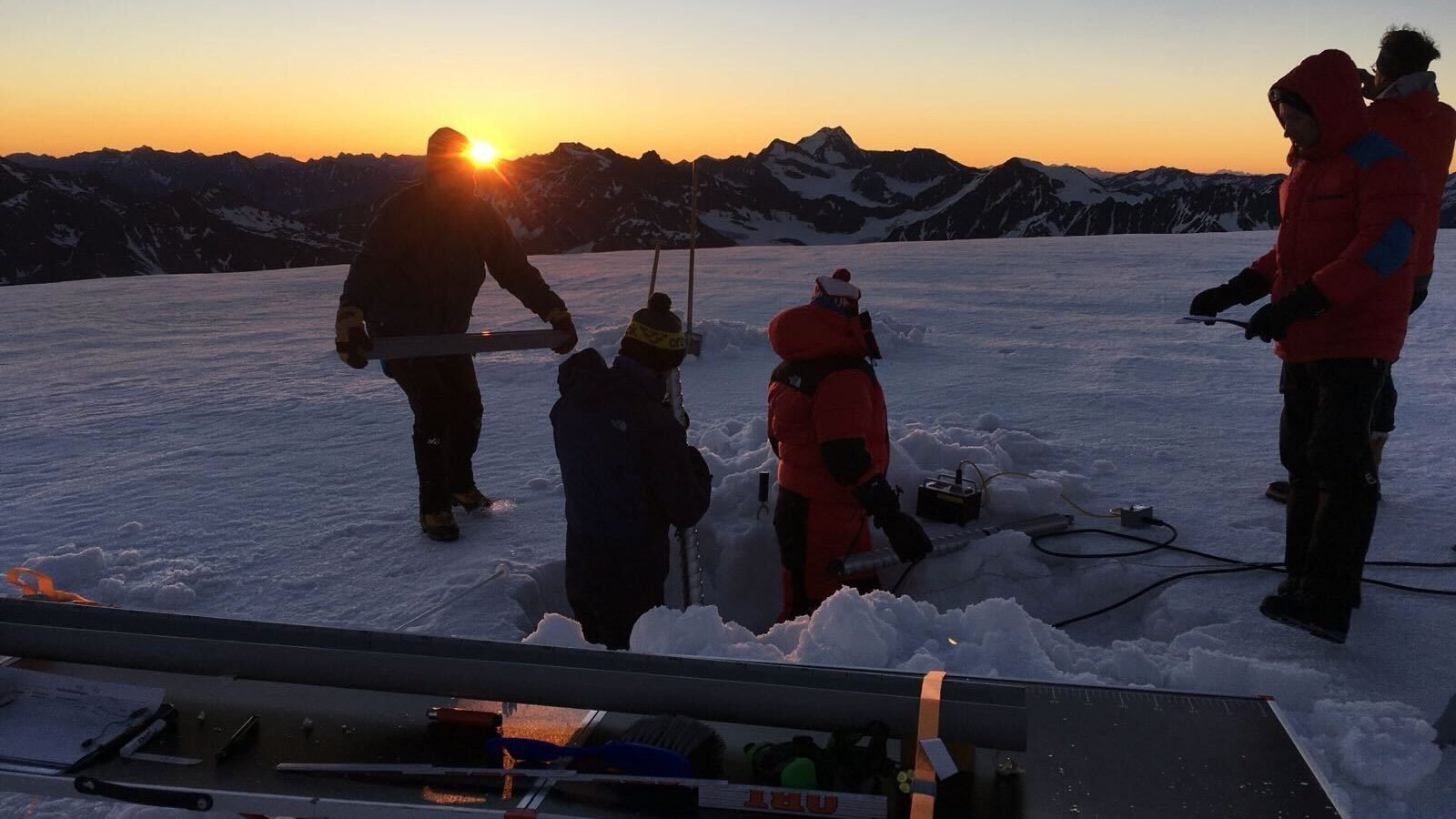 Four researchers in coats and hats stand around a hole dug in the ice on top of a glacier, while the sun peeks over the mountains behind them