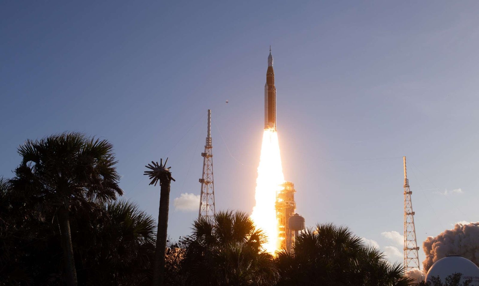 The Artemis II Moon rocket lifts off. There is a bright yellow-orange plume of fire underneath the rocket. The trees in the foreground appear in shadow.