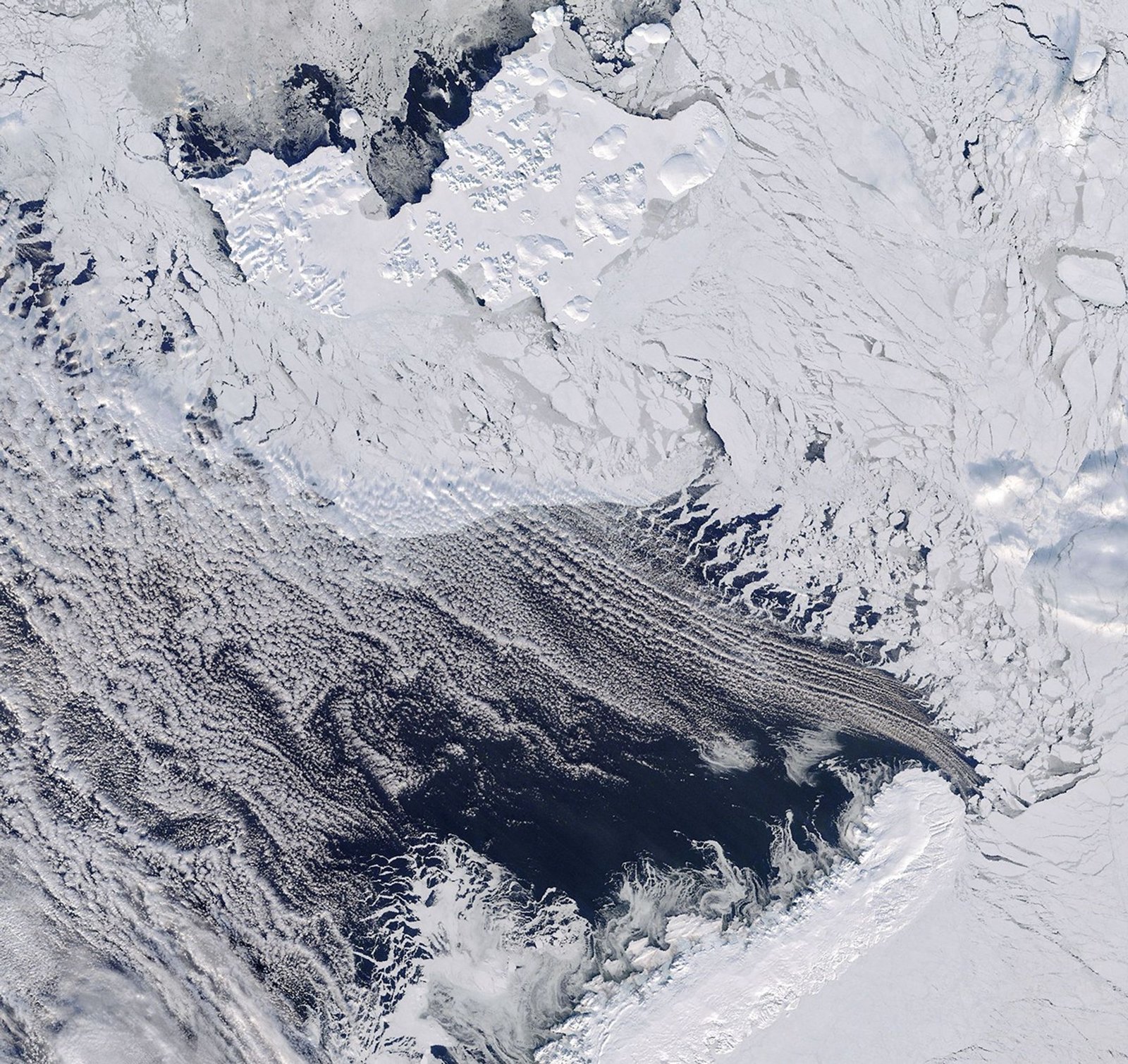 Dark open water lies south of thin, broken up sea ice near Franz Josef Land, with a thin layer of clouds covering part of the scene.