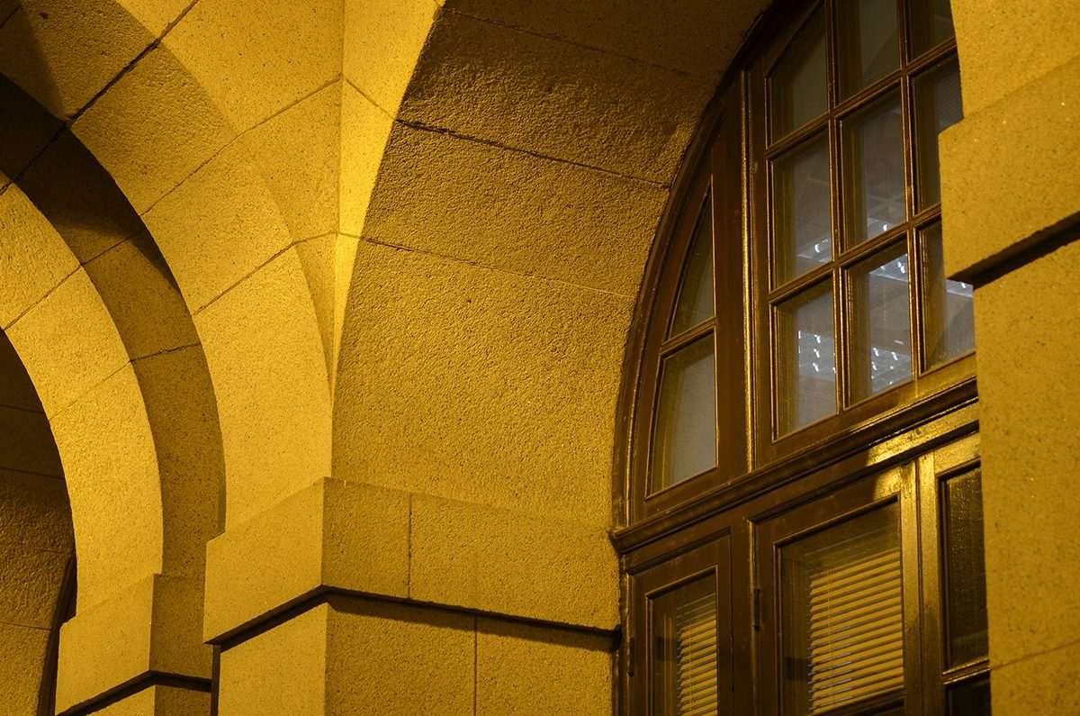 Close-up of a stone building façade with arched columns and a large wooden-framed window, illuminated by warm yellow lighting at night
