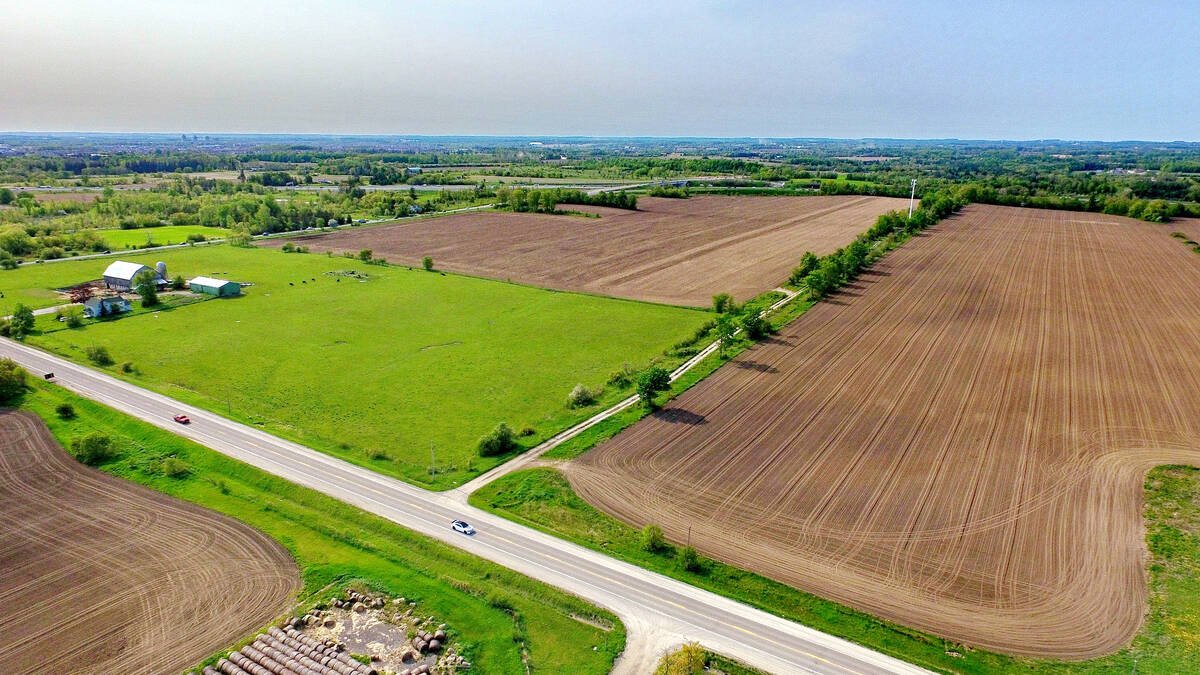 An aerial view of green grass and mostly bare agricultural cropland adjacent to a highway under partly-cloudy blue skies in southern Ontario east of Toronto. Duffins Rouge Agricultural Preserve, Durham County, Ontario, Canada. Spring 2023. Photo: Bob Hilscher/iStock/Getty Images