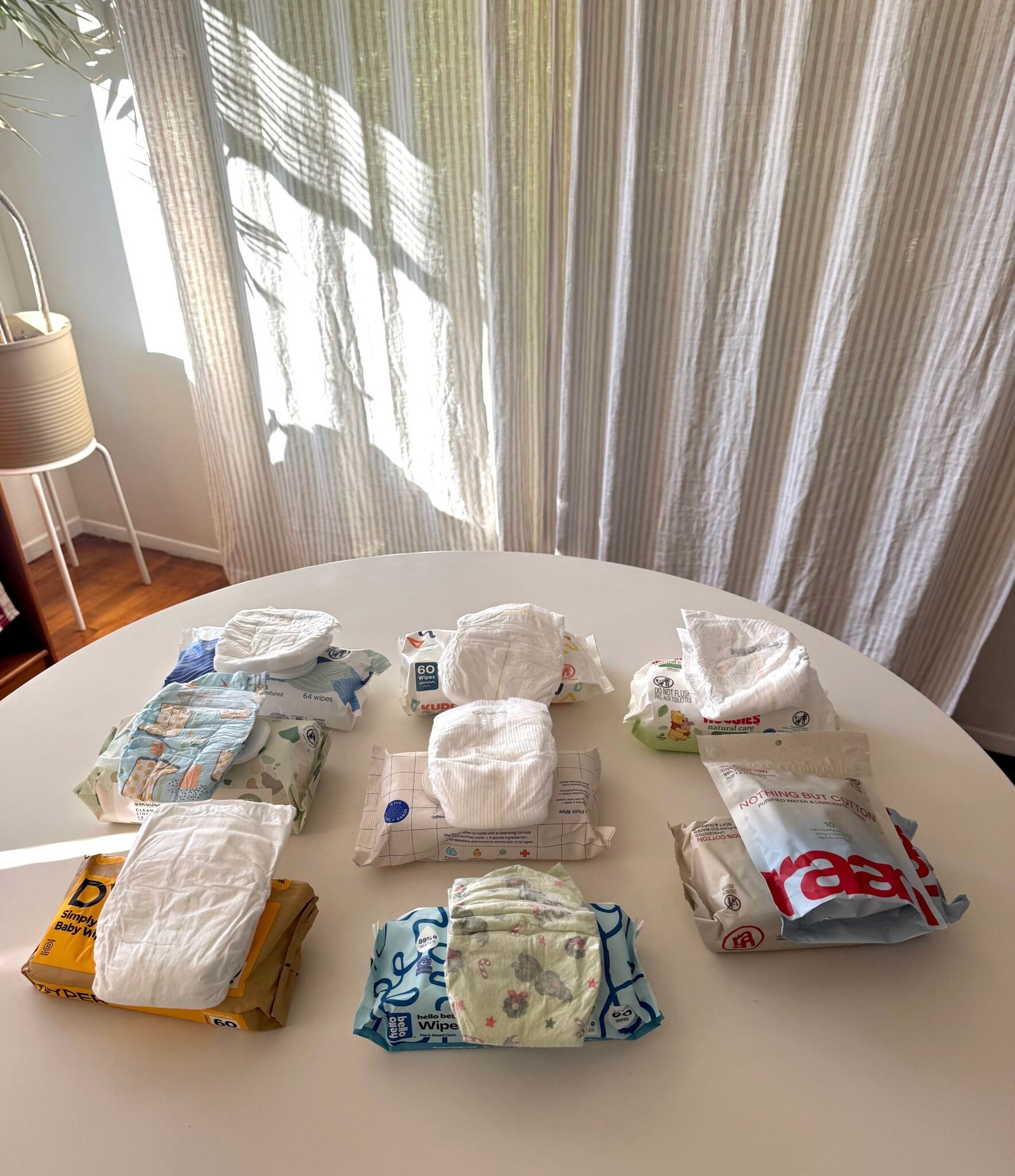 Nine different brands and types of disposable diapers are arranged in three rows on a white table in a sunlit room with sheer curtains.