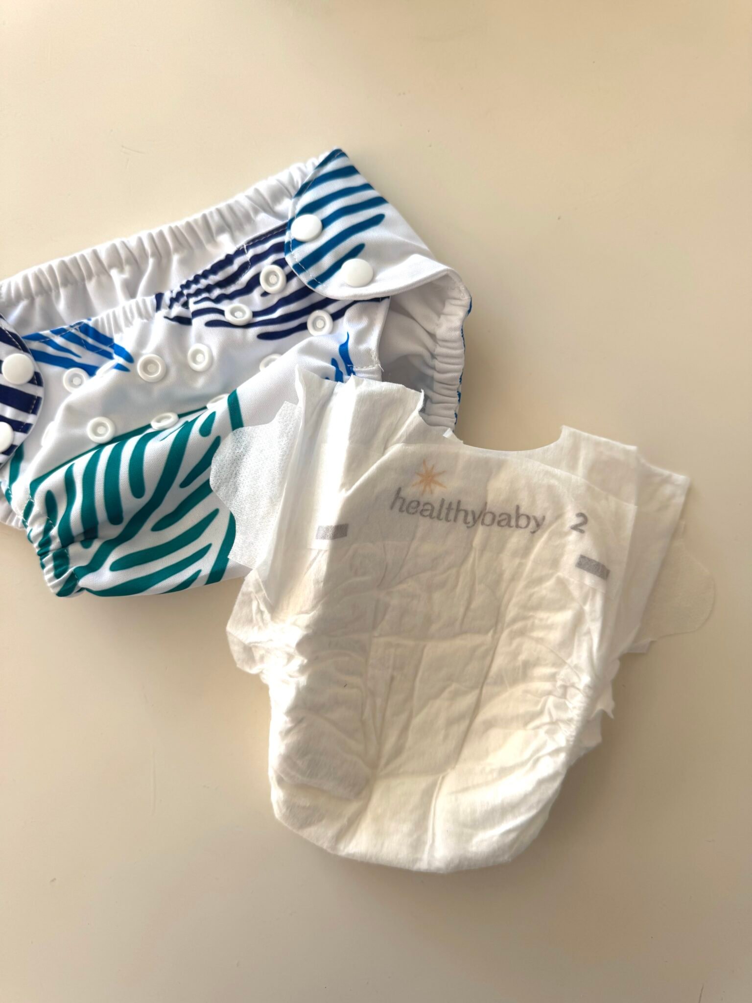 A reusable cloth diaper with blue and green stripes sits next to a disposable HealthyBaby diaper, size 2, on a white surface.