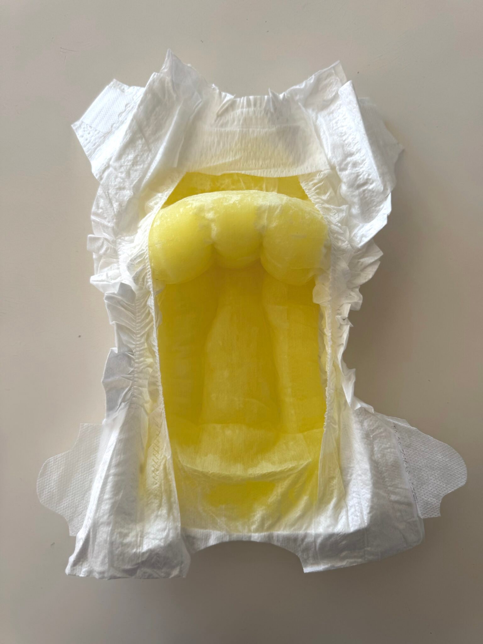 An open disposable diaper with a visible yellow liquid absorbed in the center, displayed on a neutral background.